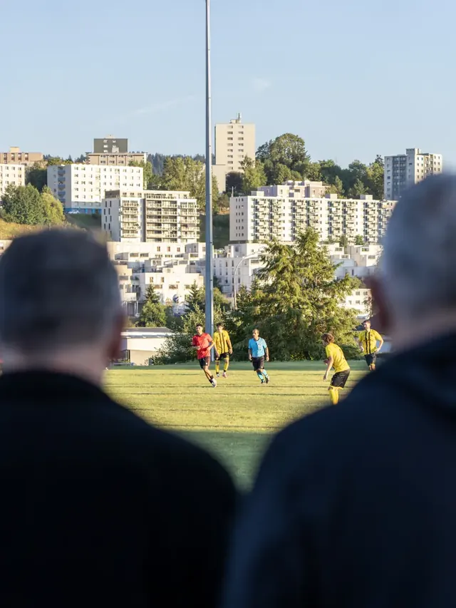 Deux spectateurs de dos regardent un match, immeubles en arrière-plan.