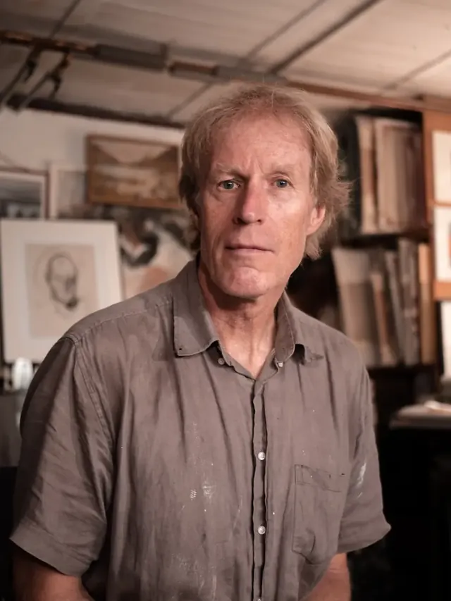Portrait d’un homme debout dans son atelier, dessins et cadres derrière lui.