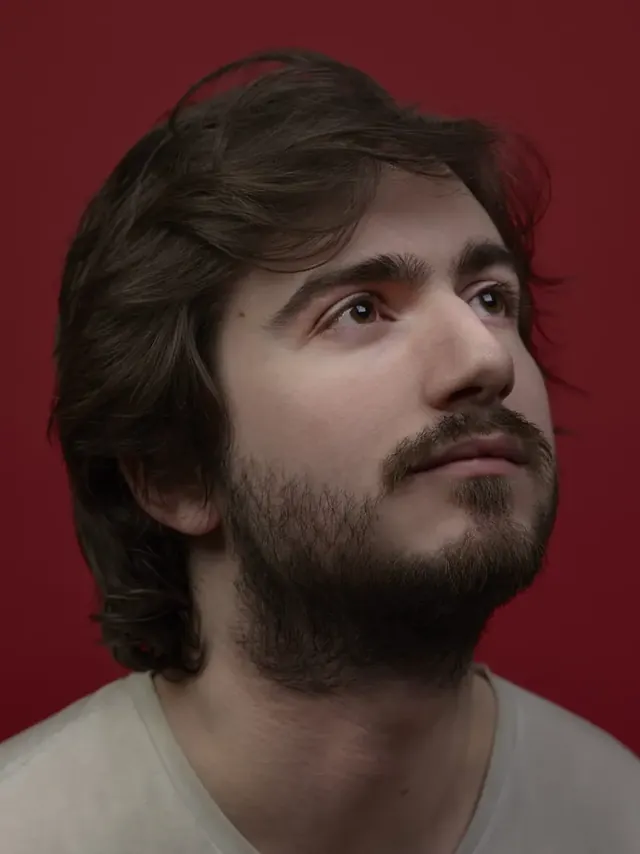 Portrait de trois quarts d’un homme barbu sur fond rouge, en studio.