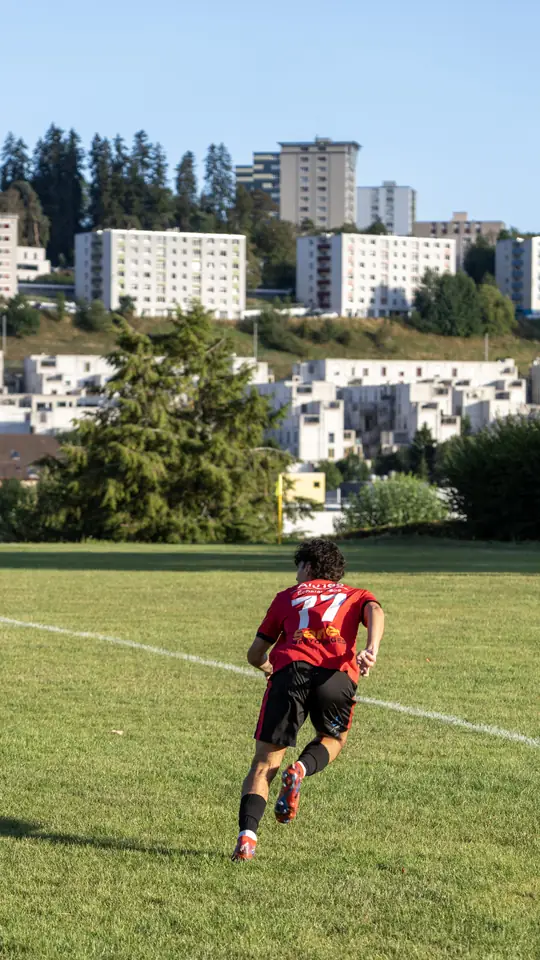 Joueur du FC Kosova en maillot rouge et noir, courant sur le terrain devant les immeubles de Neuchâtel.